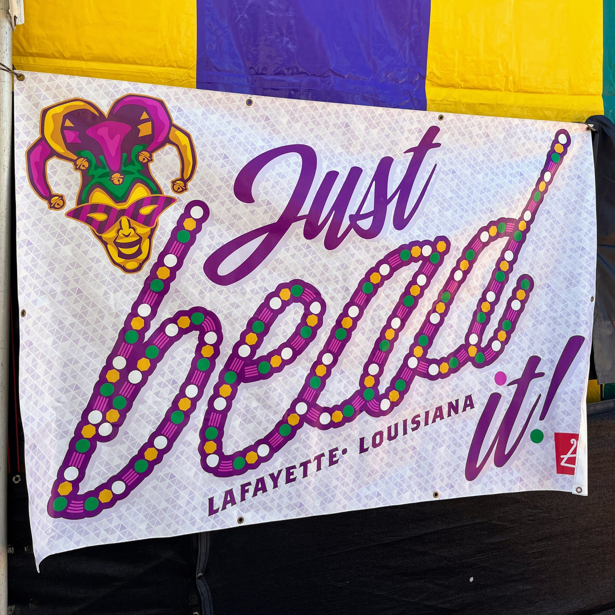 Banner reading “Just bead it! Lafayette, Louisiana,” decorated with Mardi Gras colors, beads, and a jester mask
