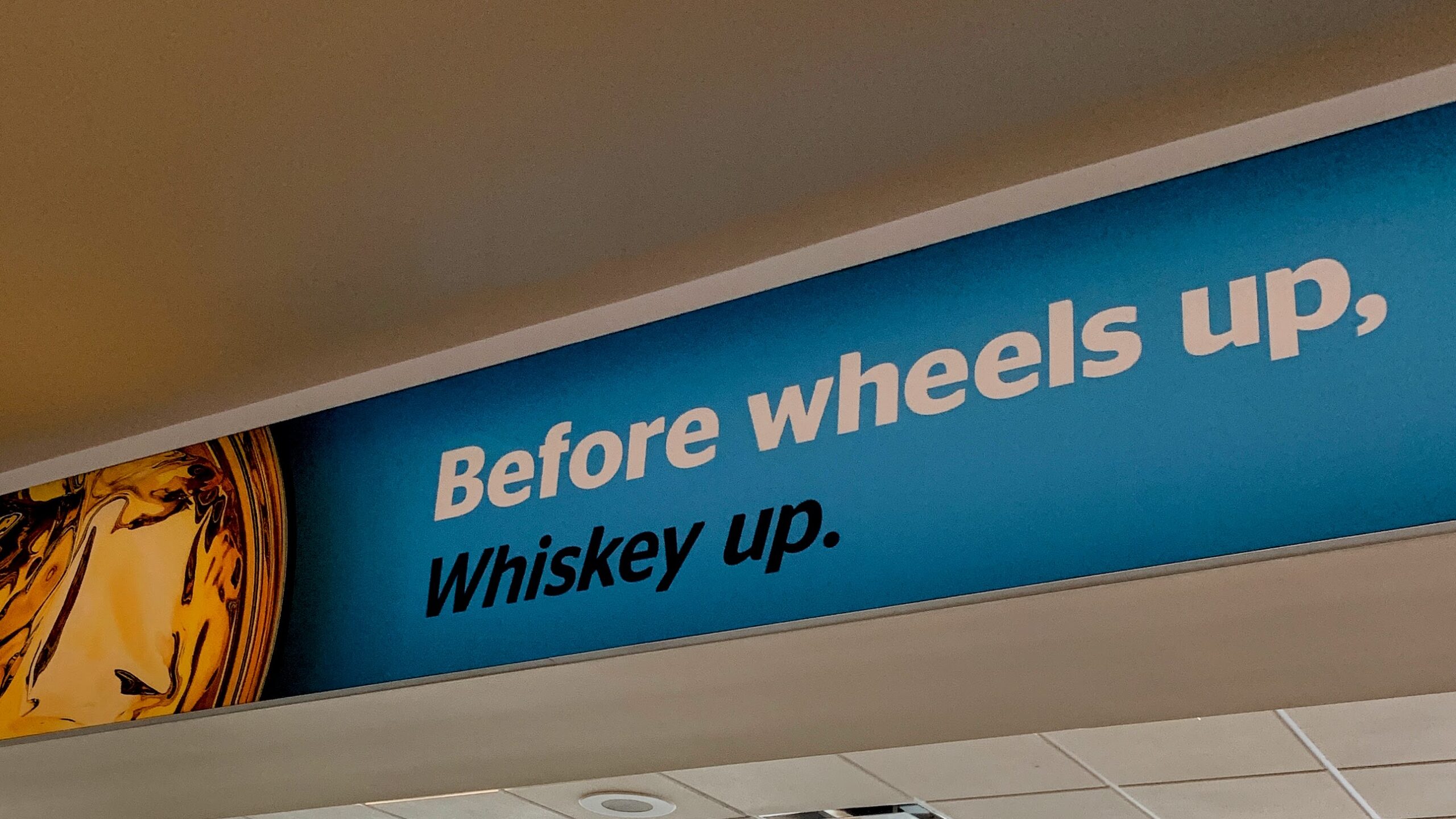 Airport sign reading “Before wheels up, Whiskey up” above a terminal walkway.